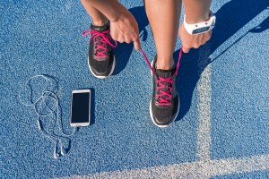 Runner woman tying running shoes laces getting ready for race on run track with smartphone and earphones for music listening on mobile phone. Athlete preparing for cardio training. Feet on ground.